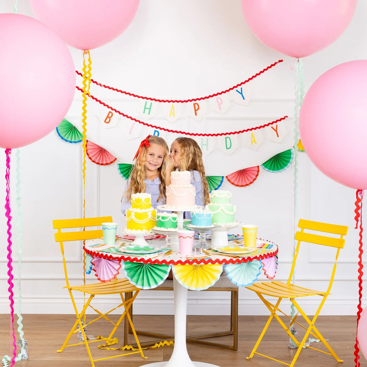Birthday dessert table with layered pastel cakes and assorted party cups in bright colors.