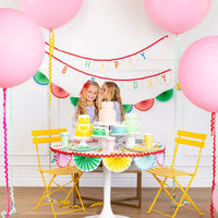 Birthday dessert table with layered pastel cakes and assorted party cups in bright colors.