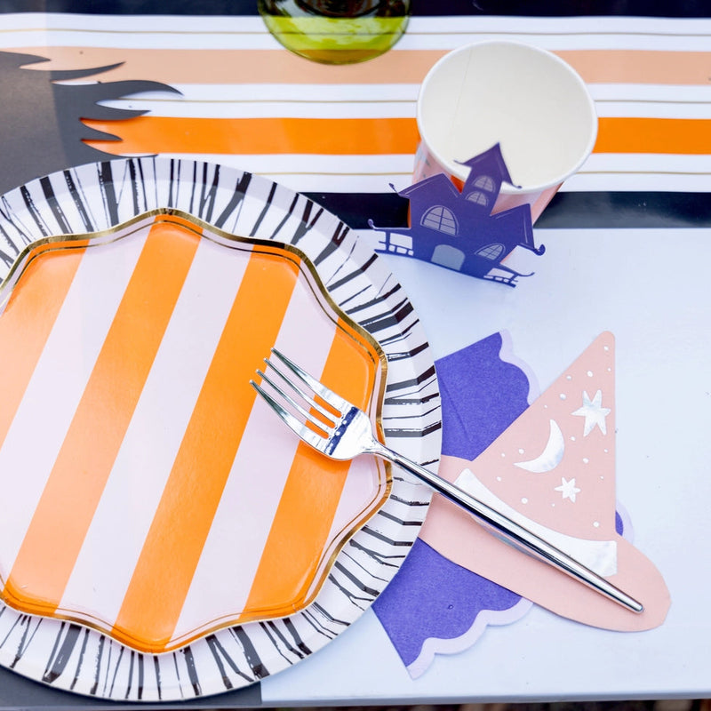 Place setting on striped runner with orange striped plate and a lilac scalloped cocktail napkin tucked by the fork.