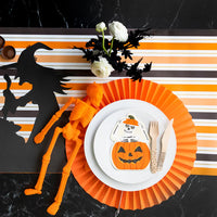 White dinner plate with skeleton pumpkin napkin on striped runner, orange skeleton decor on the side.