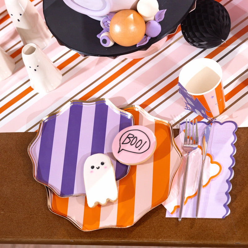 Layered purple and orange striped plates on a pink-orange striped runner with a lilac scalloped guest towel tucked under the fork and ghost and “BOO” cookies on top.