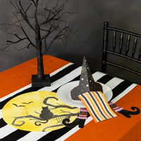 Moody table with orange cloth and black striped runner, white plate topped with hat napkin holder and striped napkin, witch’s legs accents underneath.