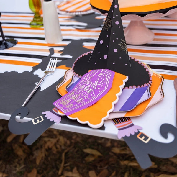 Close place setting on striped runner with stacked purple and orange plates, witch hat napkin holder, and witch’s legs accents peeking from both sides.