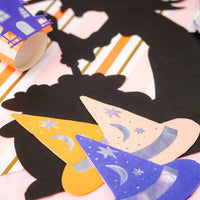 Witch silhouette placemat on a pink and orange striped runner with a purple haunted house cup and three pastel witch hat napkins.