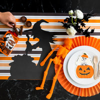 Striped runner with witch silhouette placemat beside a white plate set topped with a skeleton in pumpkin napkin and orange paper fan charger.