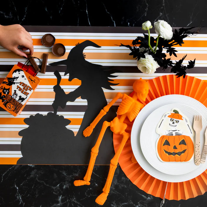 Striped runner with witch silhouette placemat beside a white plate set topped with a skeleton in pumpkin napkin and orange paper fan charger.