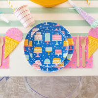 Overhead shot of stacked large and small blue cake-print plates with pink napkin, wood cutlery, and ice-cream cone napkin
