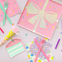 Overhead party setup with candle napkins layered under pink forks, gift-box plate, and confetti for a colorful birthday table