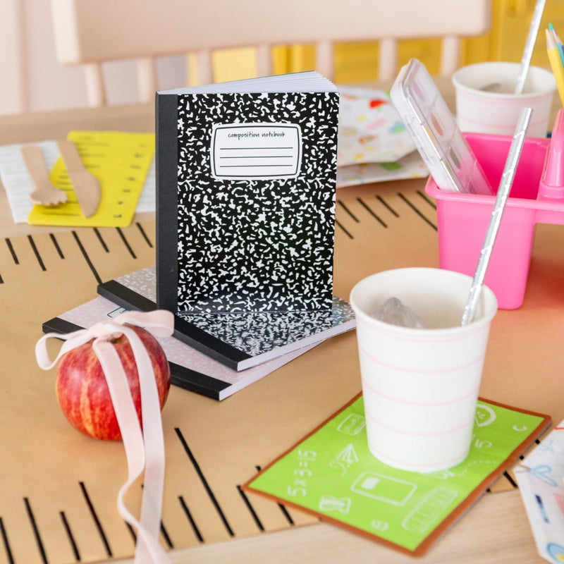 Side view of chalkboard napkin used as coaster beside composition notebook stack, apple, and cups with silver straws