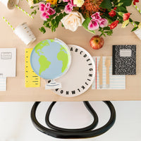 Overhead table scene showing globe plate atop alphabet charger, flowers, ruler napkin runner, and composition notebooks