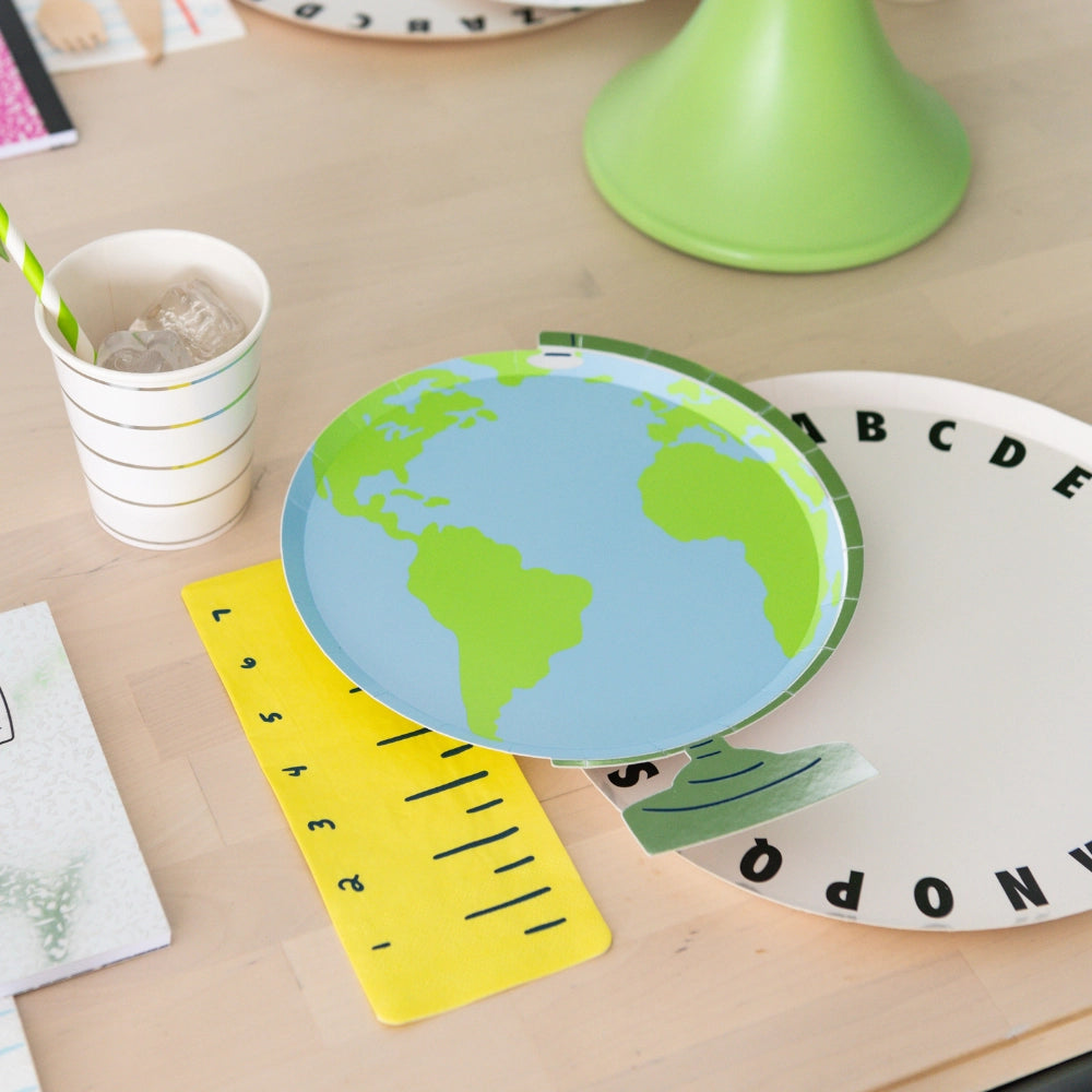 Close-up lifestyle shot of globe plate resting on yellow ruler napkin beside striped cup and green cake stand	