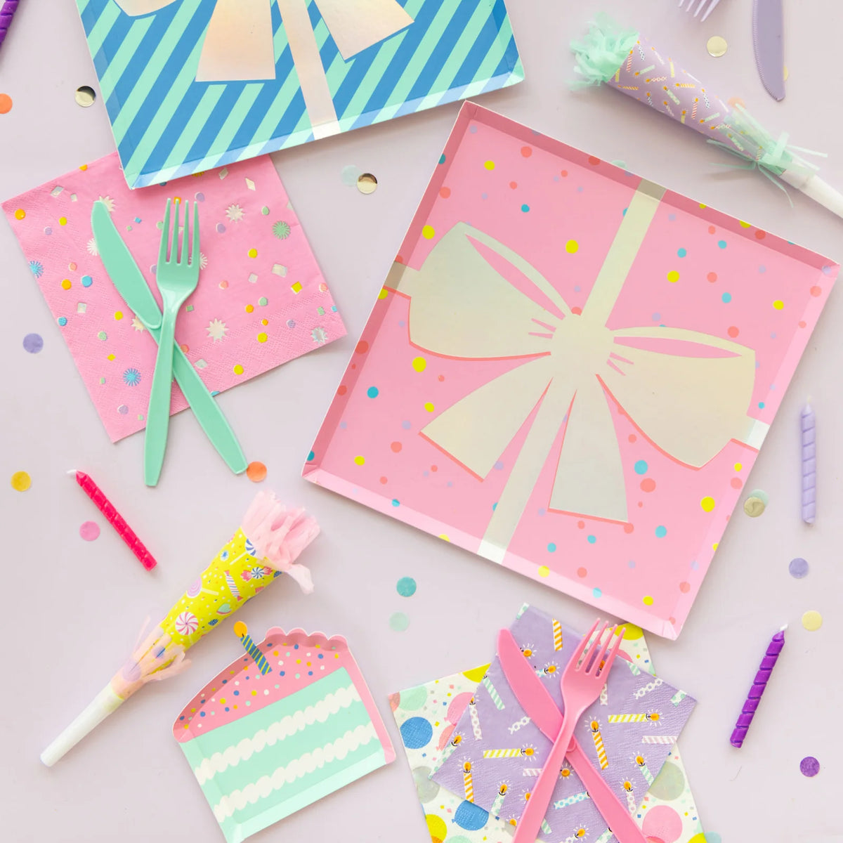 Flat-lay of birthday supplies—pink gift-box plate, candle napkin, cake-slice plate, and assorted noise makers on lavender background