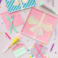Flat-lay of birthday supplies—pink gift-box plate, candle napkin, cake-slice plate, and assorted noise makers on lavender background