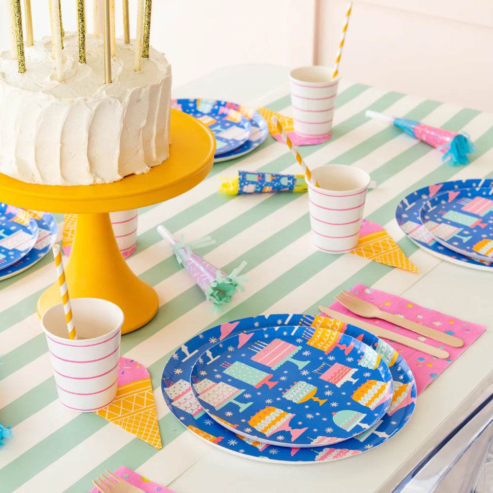 Styled birthday table with cake, striped runner, blue cake-print plates, pink napkins, and assorted noise makers