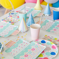 Birthday table set with cake-shaped plates, balloon plates, pastel cups, confetti, and both blue and rainbow striped party hats