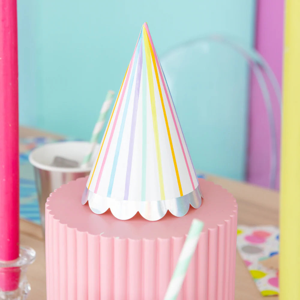 White party hat with vertical rainbow stripes and iridescent scallop edge placed on pink pedestal at a colorful birthday table