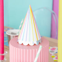 White party hat with vertical rainbow stripes and iridescent scallop edge placed on pink pedestal at a colorful birthday table