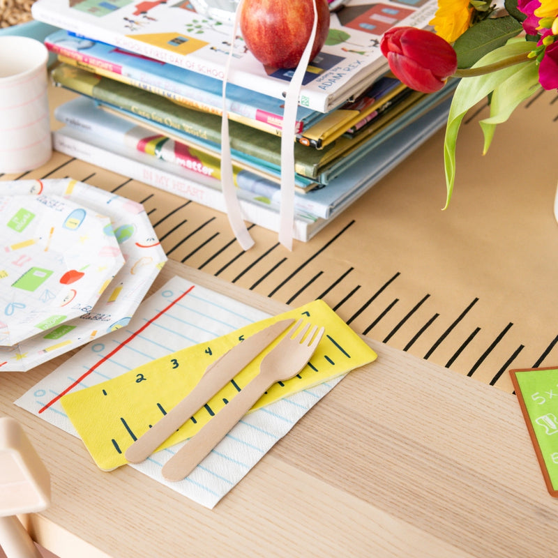 Breakfast table scene with ruler napkin atop notebook paper napkin, wooden fork and knife ready beside stacked school books and plates	