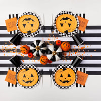 Overhead tablescape with checkerboard chargers and jack-o-lantern pumpkin paper plates, bold Halloween plates look