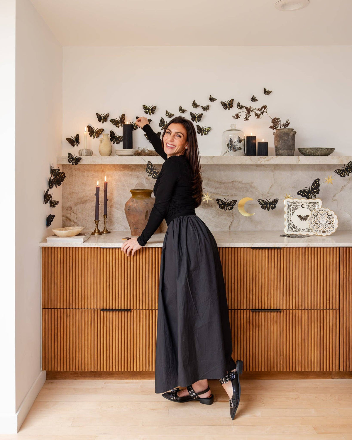 Full room shot of the Moonlight Halloween scene — woman styling moth wall decor above a wood-paneled cabinet with Moonlight plates, napkins, candles, and dark-academia decor.