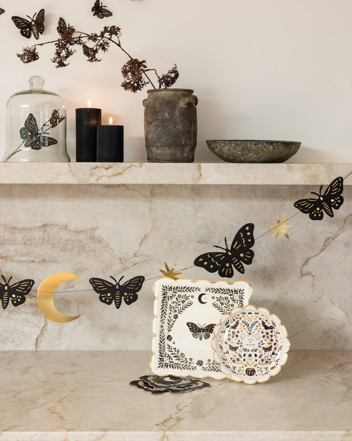 Moonlight collection shelf styling showing moth napkins beside dark candles and pottery, with black gold-foil moth wall decor on the marble wall above.