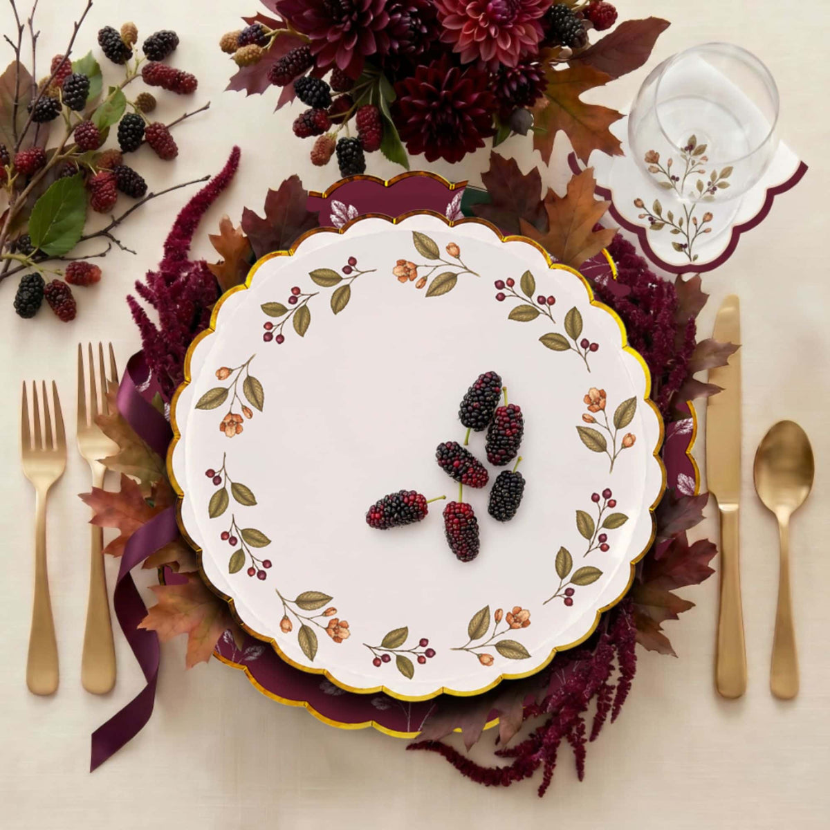 Overhead place setting with the mulberry-wreath plate on a burgundy scalloped charger, gold cutlery, and a fall floral centerpiece.