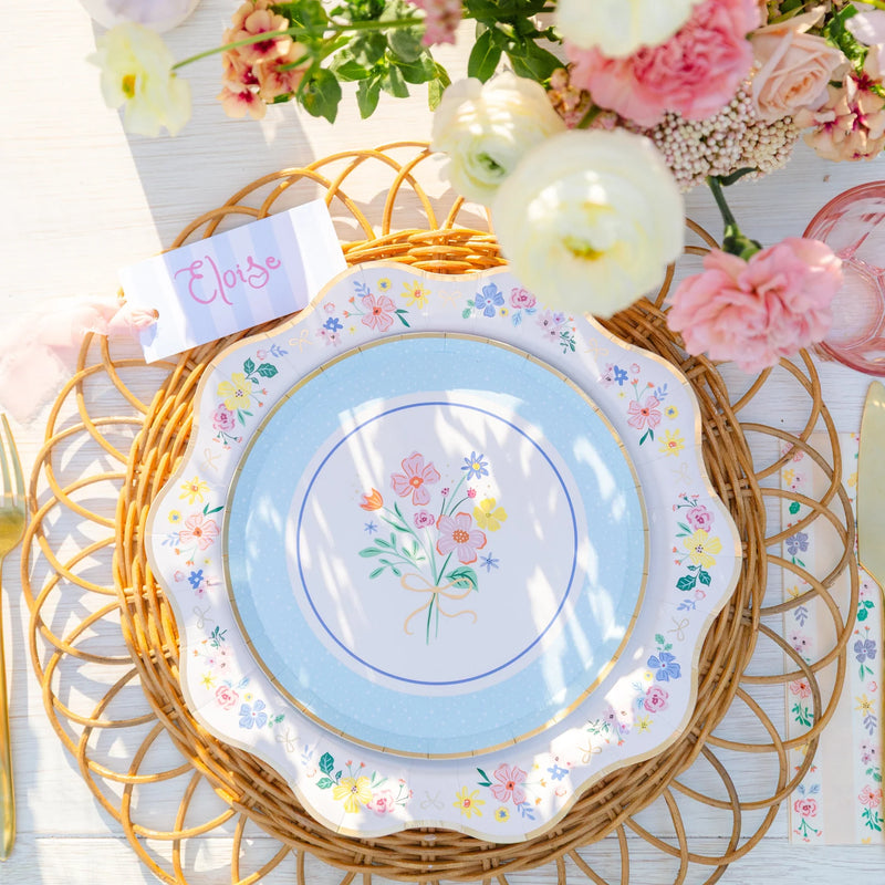 Overhead view of elegant floral scalloped dinner plate styled on a spring Easter table with pink roses, rattan charger, and place card