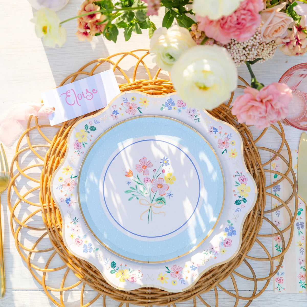 Close-up of a multicolor floral scalloped paper dessert plate with pink, yellow, and lavender floral border layered over a blue bouquet print plate on a woven rattan charger with gold flatware and a striped place card.
