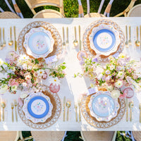 Multicolor floral scalloped paper dessert plates layered over blue floral dinner plates and rattan chargers on a spring tablescape with gold flatware, pink glassware, and garden floral centerpieces.