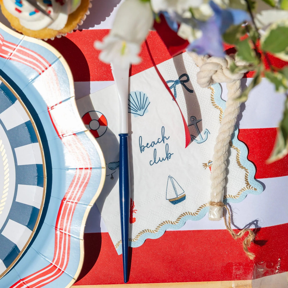 Beach club cocktail napkin styled with a nautical scalloped plate, navy fork, and rope on a red-and-white stripe runner.