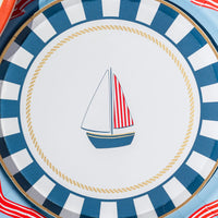 Close-up of a nautical sailboat paper dessert plate showing the navy boat, red-and-white striped sail, gold rope ring, and striped border.