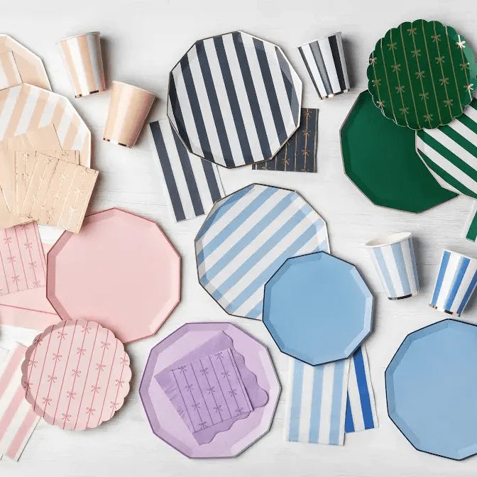 Party supplies flat lay with pink, blue, green, and lavender striped and solid paper plates, striped paper cups, and bow print napkins on a white surface.