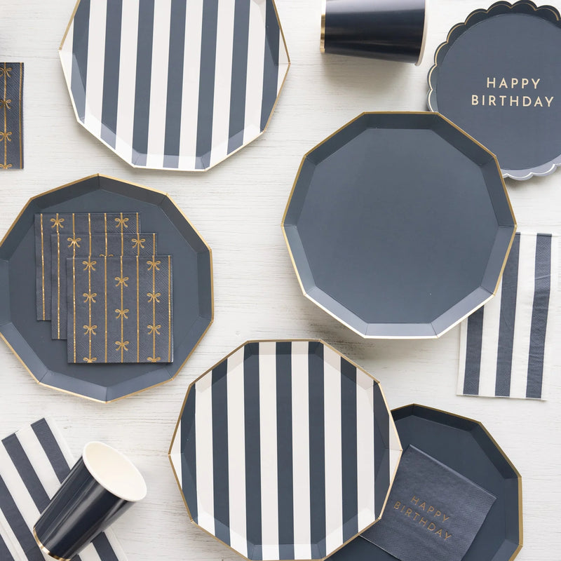 Navy and white striped and solid paper plates with a Happy Birthday plate, gold bow napkins, and navy striped guest towel napkins on a white wood surface.