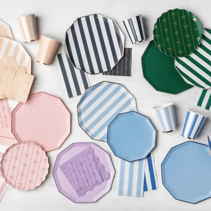 Party supplies flat lay with pink, blue, green, and lavender striped and solid paper plates, striped cups, and bow print napkins on a white surface.