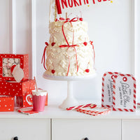 Holiday table setup showing a cake, flag banner, and the North Pole party supplies including the stacked Santa tag napkins.