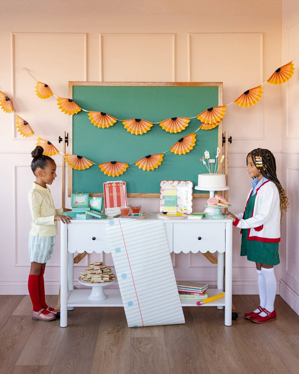 Styled back to school party with two children at white desk draped in notebook paper table runner, pencil shaving banner, chalkboard backdrop, and school-themed cake