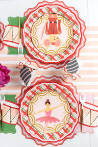 Overhead of nutcracker and ballerina plates on plaid plates with a striped ornament and a red drum napkin.