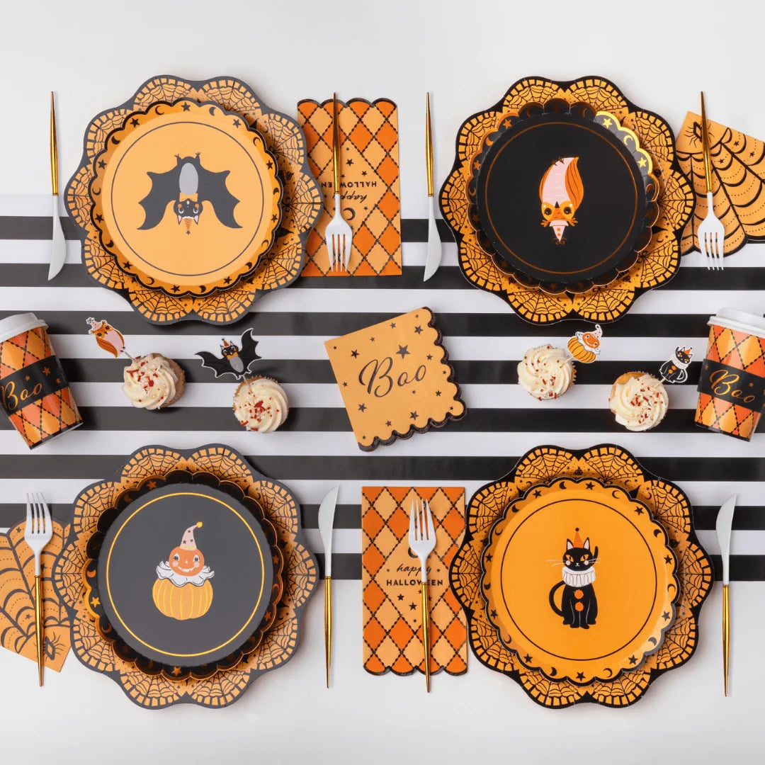 Orange argyle paper hot cups with lids and black Boo sleeves set at the ends of a vintage Halloween flat lay with character plates and cupcakes