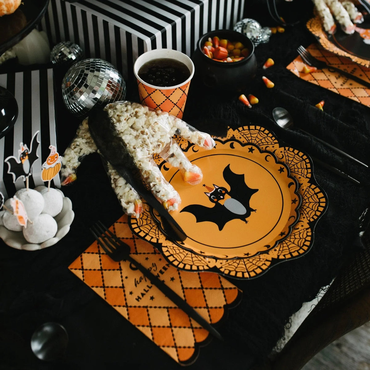 Orange argyle Happy Halloween paper guest napkin with a scalloped edge under a black fork at a kids Halloween party place setting with a bat plate and popcorn hand treat