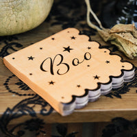 Stack of orange Boo paper cocktail napkins with black script, scattered stars, and a scalloped black edge on a black lace tablecloth beside a pumpkin