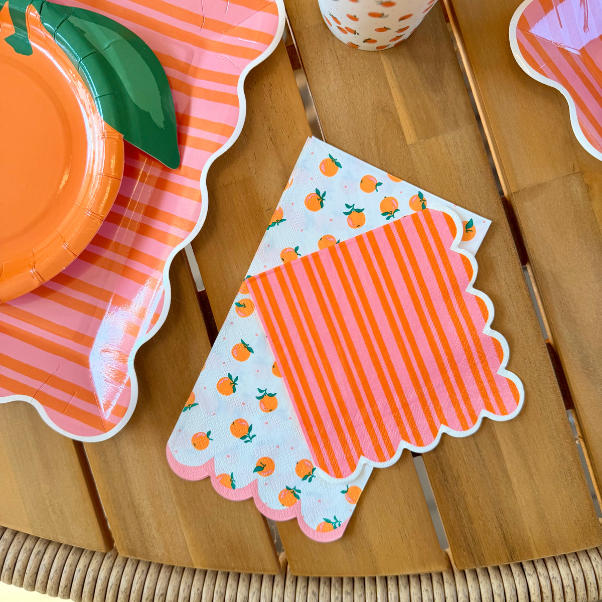 Overhead close-up of two scalloped paper cocktail napkins — orange and pink striped on top, white with scattered orange print beneath — beside a stacked set of pink striped scalloped paper plates and an orange dot paper cup