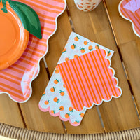 Overhead close-up of two scalloped paper cocktail napkins — orange and pink striped on top, white with scattered orange print beneath — beside a stacked set of pink striped scalloped paper plates and an orange dot paper cup