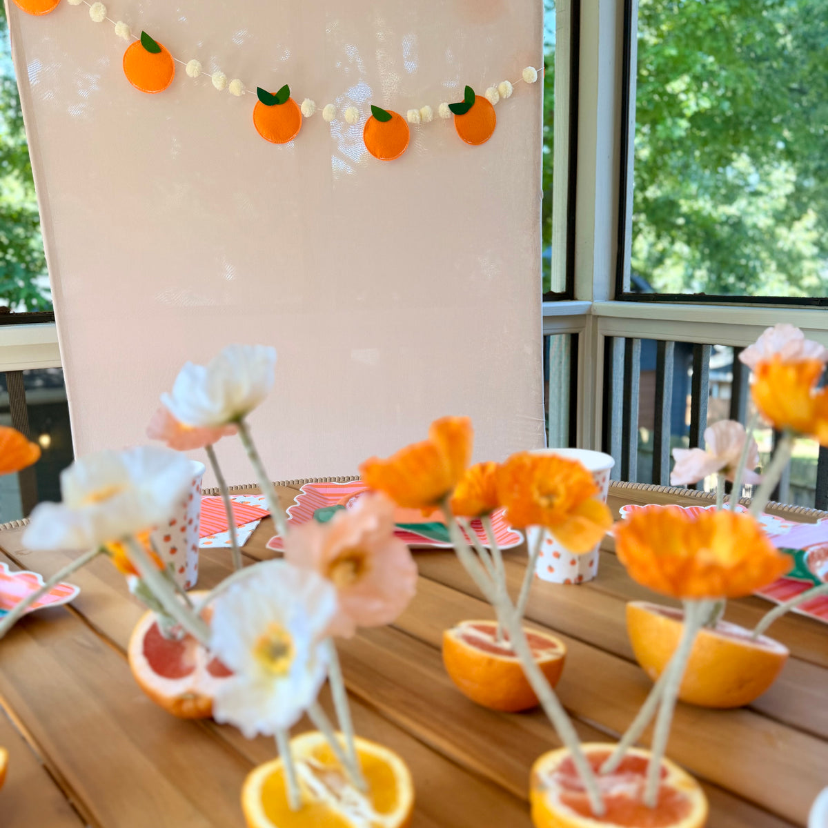 Party table styled with orange citrus paper plates and cups, set against a blush backdrop hung with a felt orange garland; halved citrus fruits holding flowers as centerpieces run the length of the table