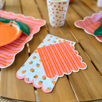  Close-up of two orange citrus scalloped cocktail napkins — one orange print on white, one orange and pink striped — displayed on a wood table alongside stacked pink striped scalloped paper plates and a white paper cup with orange dot print
