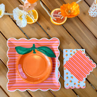 Close-up of two orange citrus scalloped cocktail napkins — one orange print on white, one orange and pink striped — displayed on a wood table alongside stacked pink striped scalloped paper plates and a white paper cup with orange dot print