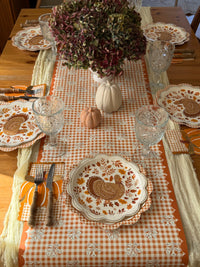 Orange plaid paper napkin with white floral sprigs and a scalloped orange edge under a pumpkin napkin and wood handled flatware, beside a turkey plate