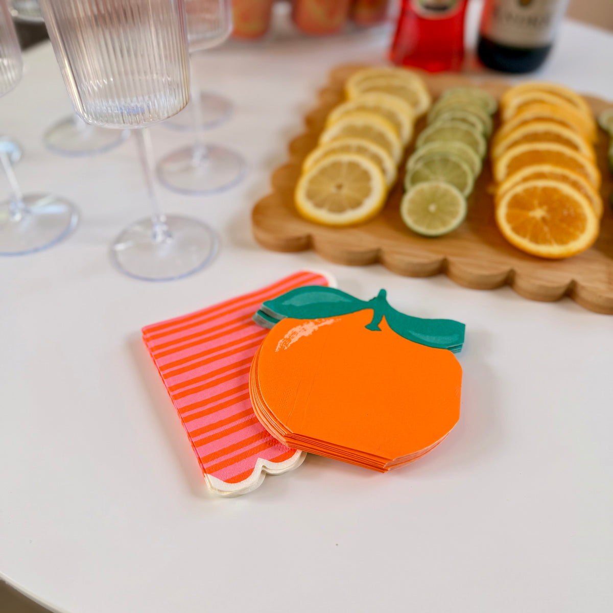 Stack of orange-shaped die-cut paper cocktail napkins beside a stack of pink and orange striped scalloped cocktail napkins, displayed on a white surface next to a citrus garnish board and champagne flutes