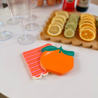 Stack of orange-shaped die-cut paper cocktail napkins beside a stack of pink and orange striped scalloped cocktail napkins, displayed on a white surface next to a citrus garnish board and champagne flutes
