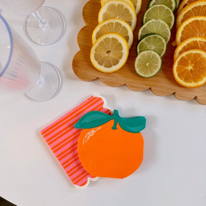 Orange-shaped die-cut paper cocktail napkins stacked with pink and orange striped scalloped napkins on a white surface, with a sliced citrus garnish tray and coupe glasses in the background