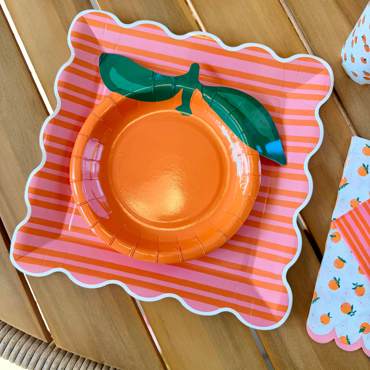 Orange-shaped paper dessert plate with green leaf detail centered on a pink and orange striped scalloped square paper dinner plate, with orange citrus napkins visible at the edge of the frame